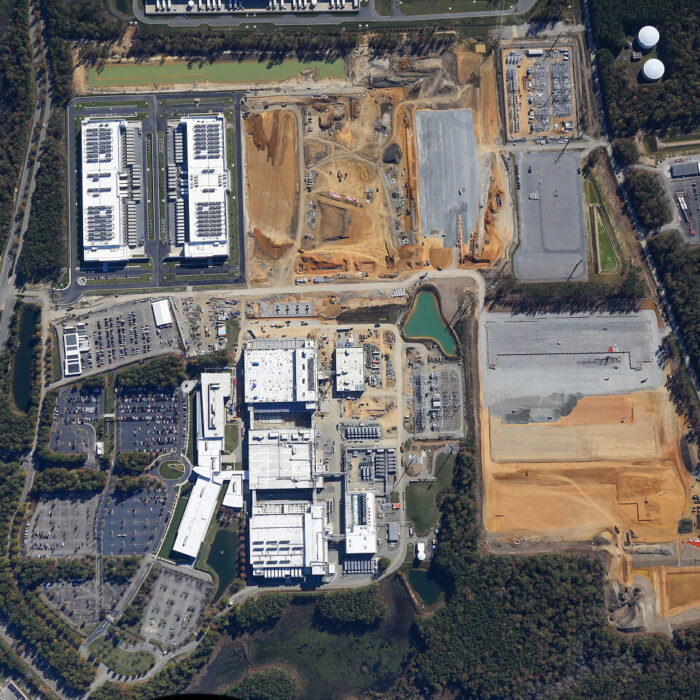 Aerial view: construction site, finished buildings, parking, roads, trees.