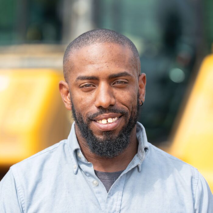 Bearded man in blue shirt stands by yellow construction equipment.