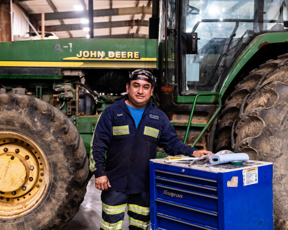 Man stands by blue tool chest and John Deere tractor indoors.