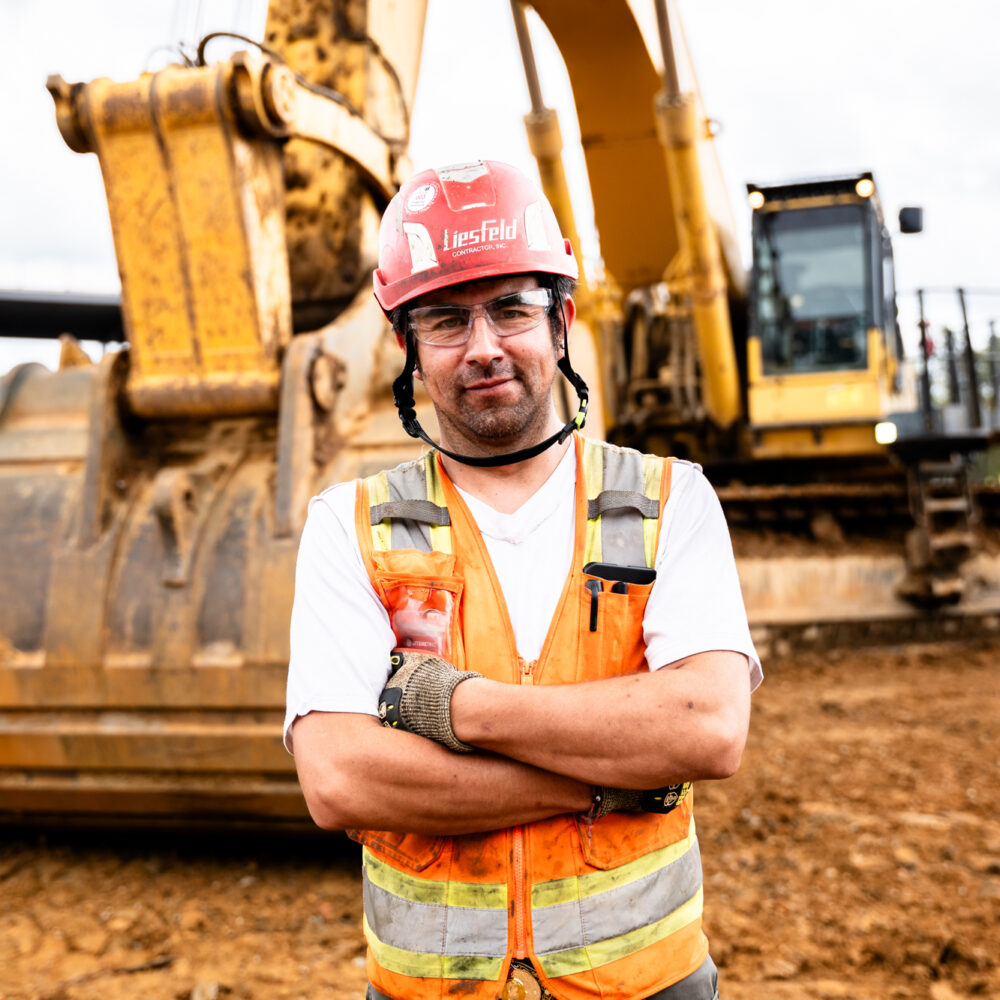 Worker in vest and helmet stands by excavator, arms crossed.